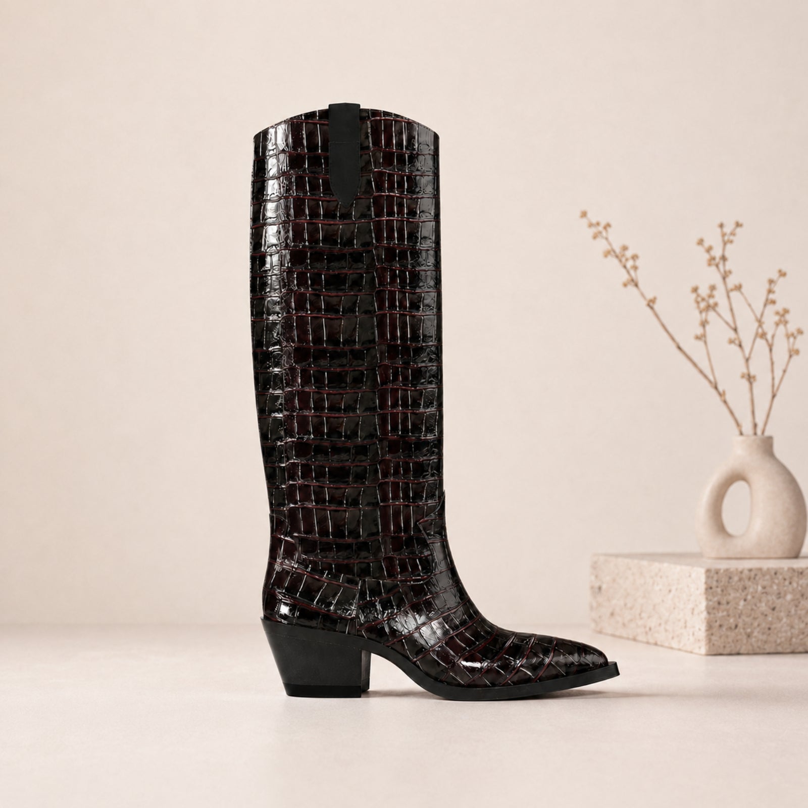 A single knee-high boot with a glossy crocodile-embossed texture in black, standing on a light beige surface next to a small decorative vase with dried stems.