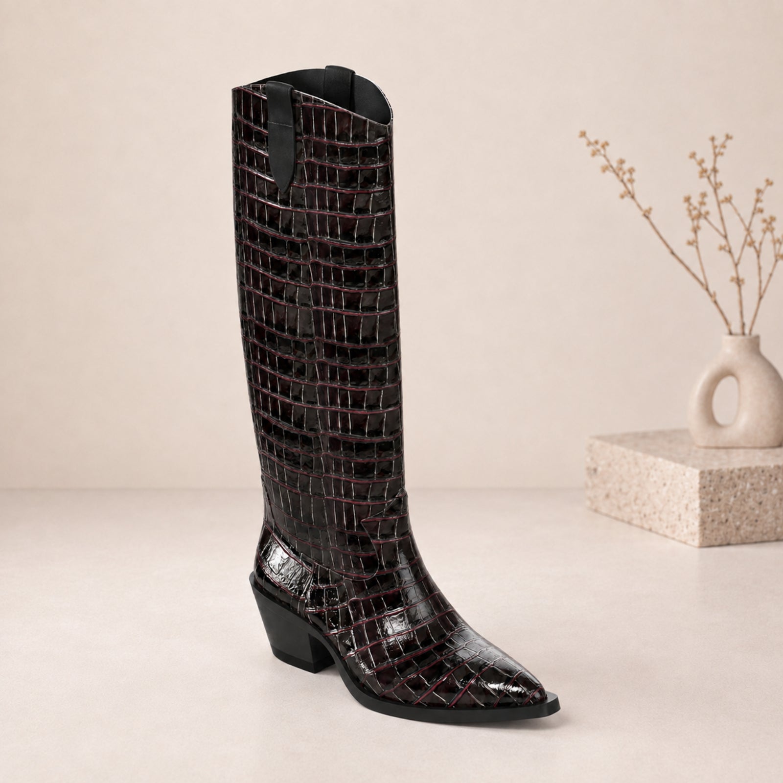 A single knee-high boot with a glossy crocodile-embossed texture in black and deep burgundy, standing on a light beige surface next to a small vase with dried branches.