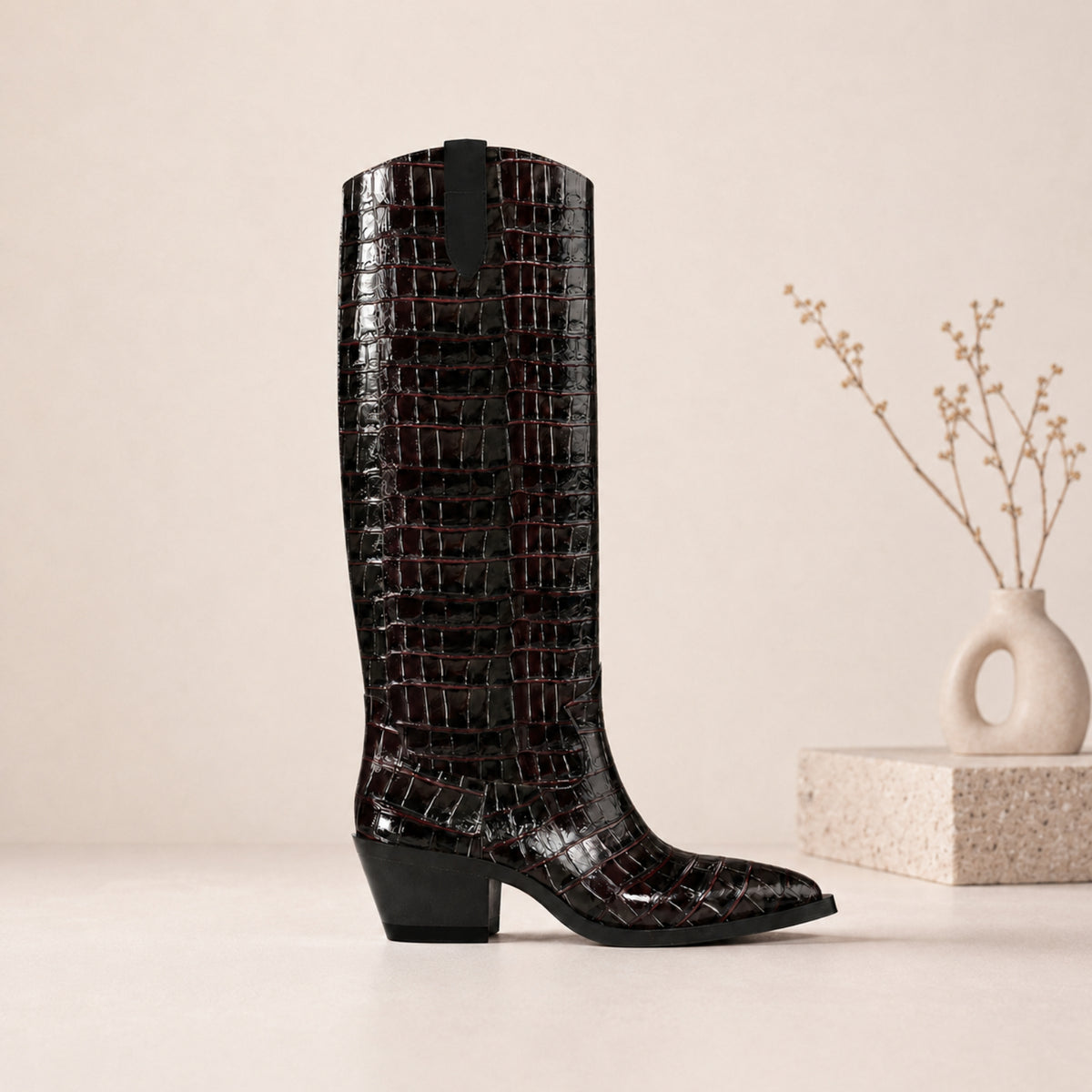 A single knee-high boot with a glossy crocodile-embossed texture in black, standing on a light beige surface next to a small decorative vase with dried stems.